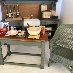 May include: A miniature dolls' house kitchen scene featuring a green wooden table with a mixing bowl, flour, raisins, and small pastries. Beside the table sits a green wicker chair. In the background, a green kitchen unit includes a sink, bread bin, and hanging utensils.
