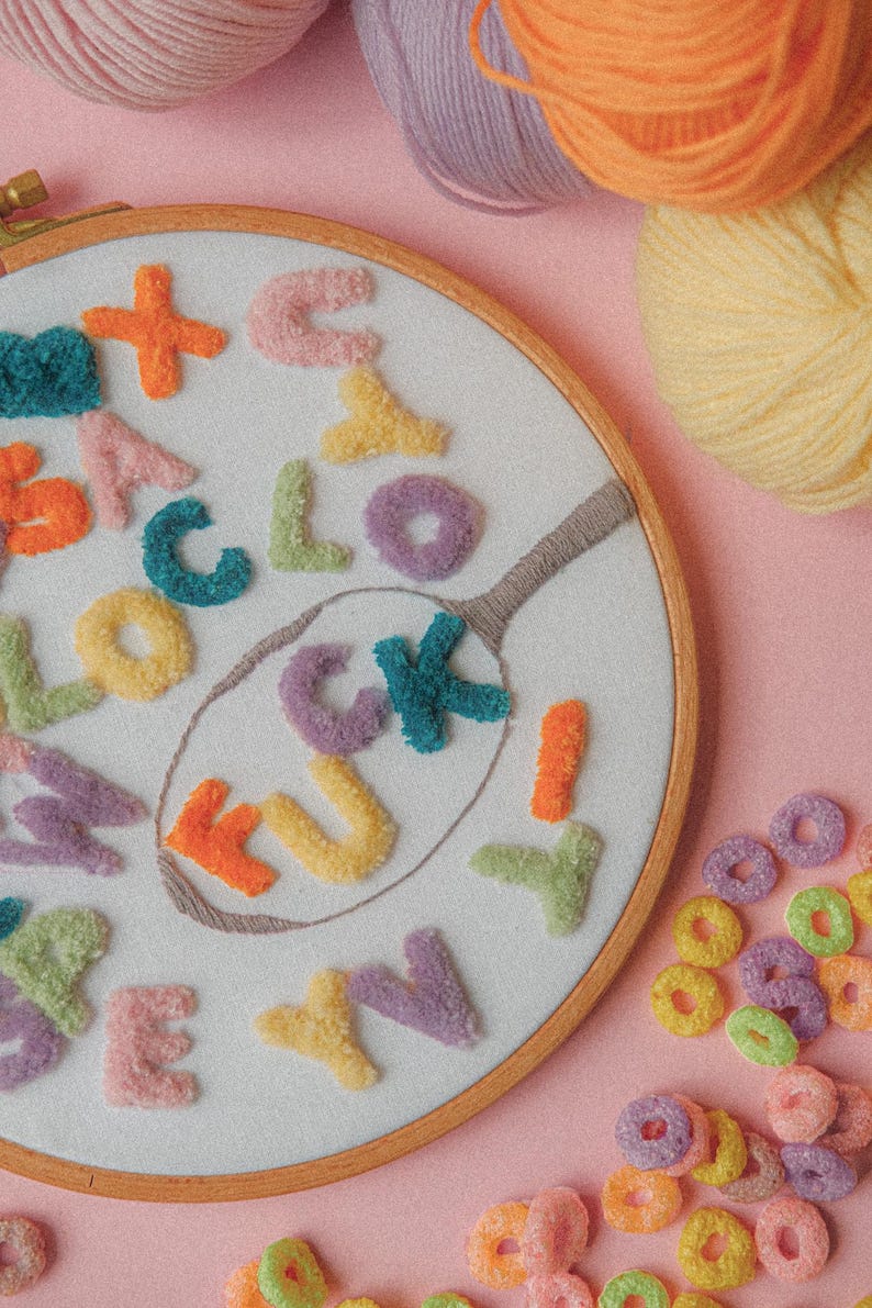 Pode incluir: Um aro de bordado branco com a palavra "F*CK" escrita em letras felpudas coloridas. As letras s&atilde;o cercadas por outras letras de v&aacute;rias cores. O aro est&aacute; sobre uma superf&iacute;cie rosa com um monte de an&eacute;is de cereal coloridos.
