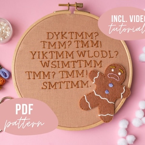 May include: An embroidery hoop with brown text and a felt gingerbread man on a pink background. The text reads "DYKTMM? TMM? TMM! YIKTMM WLODL? WSIMTTMM TMM? TMM! SMTTMM." Includes video tutorials and a PDF pattern.