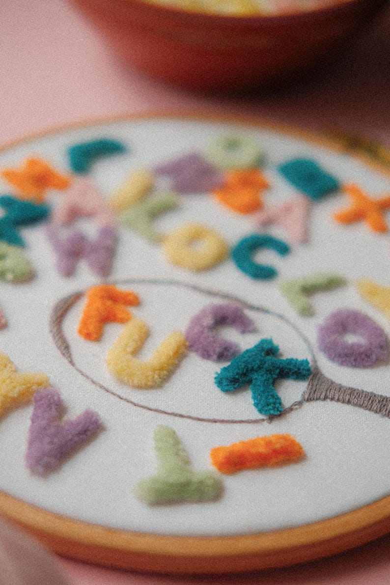 Pode incluir: Um aro de bordado branco com um design de feltro colorido da palavra "F*CK" dentro de um espelho. As letras s&atilde;o feitas de diferentes cores de feltro, incluindo laranja, amarelo, roxo e turquesa.