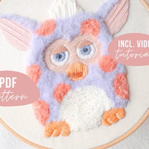 May include: A purple and pink embroidered plush toy in the shape of a fuzzy creature with large eyes. The toy is in a wooden embroidery hoop. The text "INCL. VIDEO tutorials" is on a pink rectangle in the top right corner of the image. The text "PDF pattern" is on a pink rectangle in the bottom left corner of the image.