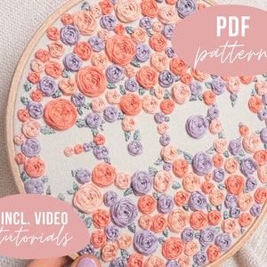 May include: A circular embroidery hoop with a floral design featuring pink and purple flowers. The hoop is on a light beige surface. The text "PDF pattern" is in a pink circle and the text "INCL. VIDEO tutorials" is in a pink rectangle.