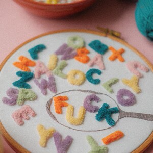 Pode incluir: Um aro de bordado branco com um design de feltro colorido da palavra "F*CK" em uma colher. As letras s&atilde;o cercadas por outras letras de feltro coloridas.
