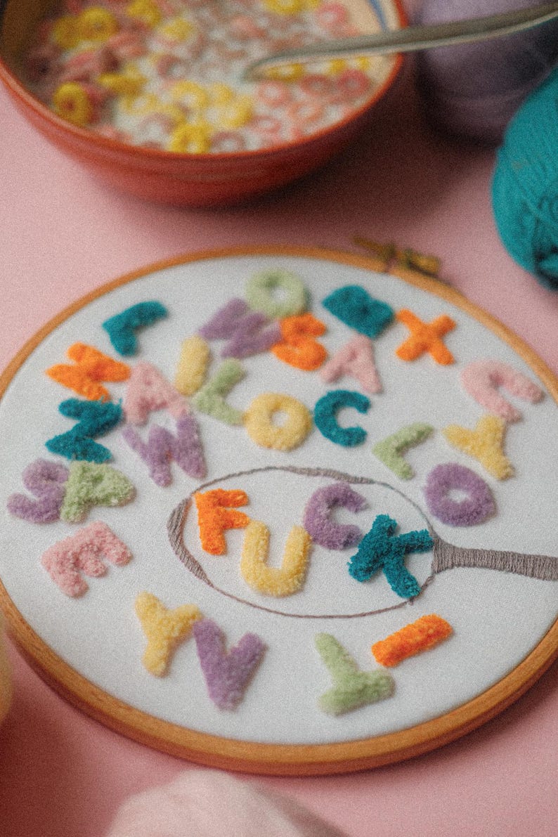 Pode incluir: Um aro de bordado branco com um design de feltro colorido da palavra "F*CK" em uma colher. As letras s&atilde;o cercadas por outras letras de feltro coloridas.