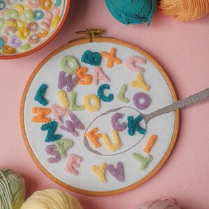 Pode incluir: Um aro de bordado branco com a palavra "F**K" escrita em letras de fio de cores diferentes. As letras s&atilde;o cercadas por outras letras de fio de v&aacute;rias cores. H&aacute; novelos de fio de diferentes cores sobre uma superf&iacute;cie rosa.