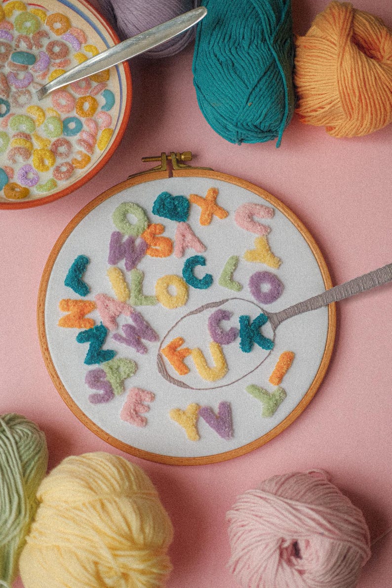 Pode incluir: Um aro de bordado branco com a palavra "F**K" escrita em letras de fio de cores diferentes. As letras s&atilde;o cercadas por outras letras de fio de v&aacute;rias cores. H&aacute; novelos de fio de diferentes cores sobre uma superf&iacute;cie rosa.