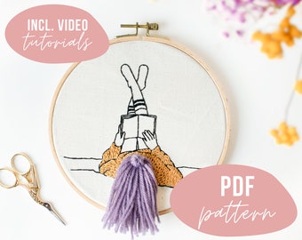 May include: Embroidery hoop art featuring a person reading a book with their legs up. The design is stitched in black and gold thread on a white fabric. The text "INCL. VIDEO TUTORIALS" and "PDF pattern" are included.