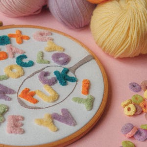 Pode incluir: Um close-up de um aro de bordado branco com a palavra "LUCKY" escrita em letras de fio de l&atilde; coloridas. O aro &eacute; cercado por novelos de l&atilde; coloridos e an&eacute;is de cereal coloridos.