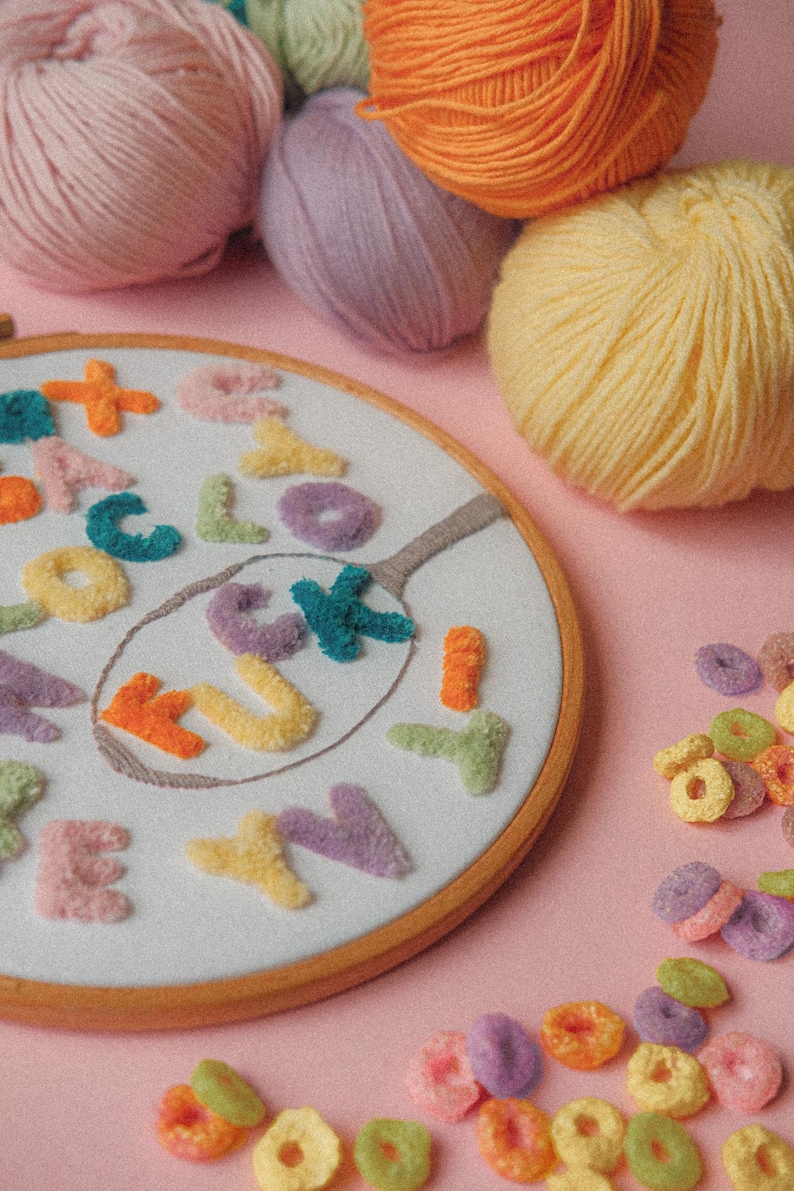 Pode incluir: Um close-up de um aro de bordado branco com a palavra "LUCKY" escrita em letras de fio de l&atilde; coloridas. O aro &eacute; cercado por novelos de l&atilde; coloridos e an&eacute;is de cereal coloridos.