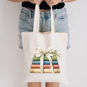 May include: A cream-colored canvas tote bag featuring a graphic print of a stack of four vintage-style books in green, blue, pink, and beige. The books are tied together with a large, decorative cream ribbon with a delicate leaf pattern.