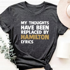 May include: A dark grey t-shirt with the text "My thoughts have been replaced by Hamilton lyrics" in white and gold lettering.