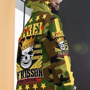 May include: A camouflage-patterned hoodie with a skull graphic, crown, and the words "REBEL" and "LIGHT DEFEATS DARKNESS." The hoodie has yellow star accents and a yellow stripe down the sleeve. The brand "XFRISSON MICHAEL SEAN JEANS" is also visible.