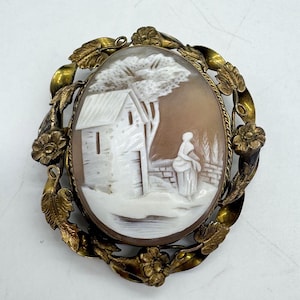 May include: Oval cameo brooch with a detailed relief carving of a rural scene. The cameo features a white building, tree, and figure against a brown background. The cameo is set in a gold-toned metal frame with floral and leaf accents.