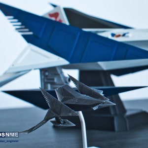 May include: A black and gray 3D printed model of a spaceship with sharp angles and a pointed nose. The spaceship is on a stand and is in front of a larger, blue and white spaceship.