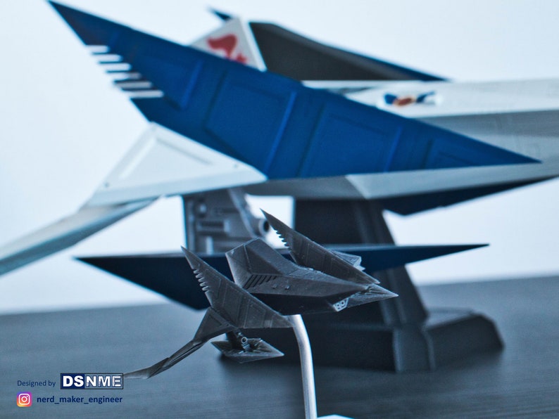 May include: A black and gray 3D printed model of a spaceship with sharp angles and a pointed nose. The spaceship is on a stand and is in front of a larger, blue and white spaceship.