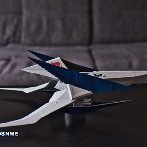 May include: A white and blue model spaceship with a red and white emblem on the side. The spaceship has a sharp, angular design with multiple wings and a pointed nose.