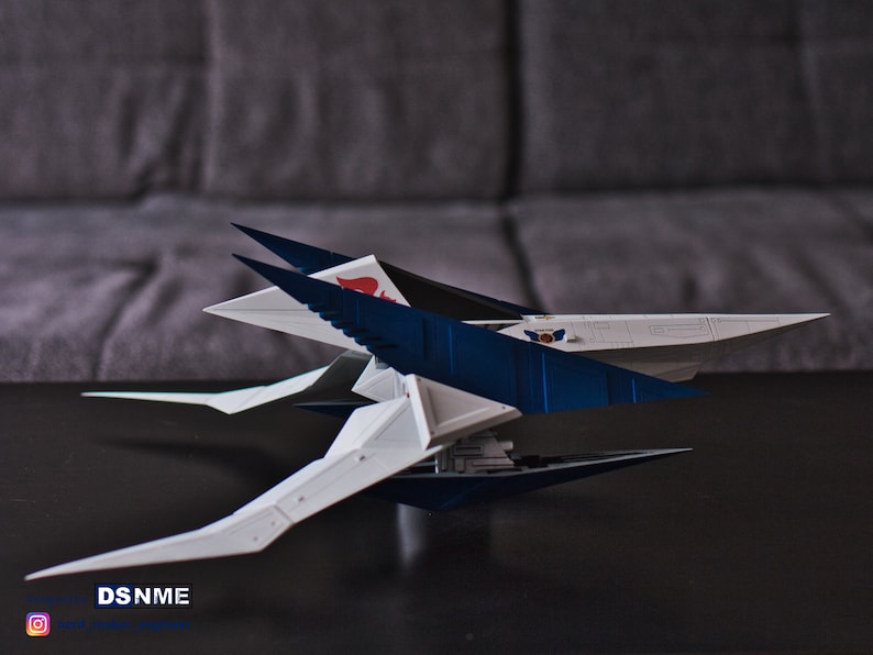 May include: A white and blue model spaceship with a red and white emblem on the side. The spaceship has a sharp, angular design with multiple wings and a pointed nose.