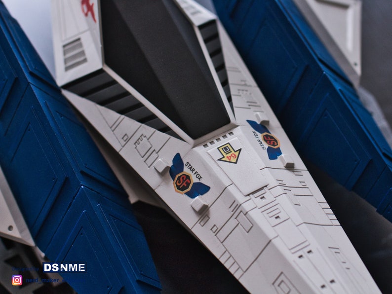 May include: A white and blue model spaceship with the words "Star Fox" and "SF" printed on the side. The spaceship has a black cockpit and a blue wing.