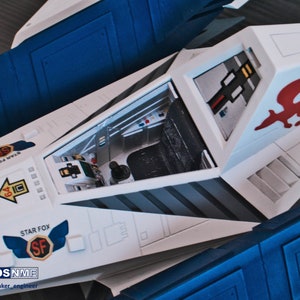 May include: A white and blue model of a Star Fox spaceship with an open cockpit. The cockpit has a black seat and a control panel. The spaceship has a red and white fox logo on the side. The text "STAR FOX" is printed on the side of the spaceship.