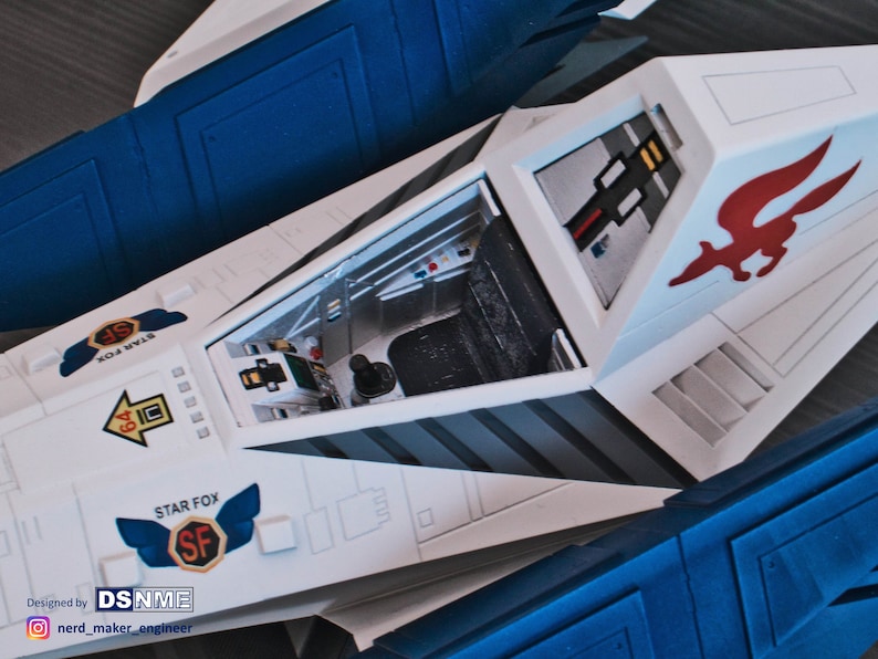 May include: A white and blue model of a Star Fox spaceship with an open cockpit. The cockpit has a black seat and a control panel. The spaceship has a red and white fox logo on the side. The text "STAR FOX" is printed on the side of the spaceship.