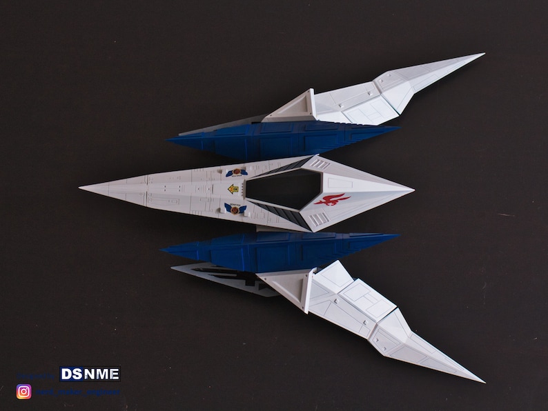 May include: A white and blue model spaceship with red and yellow accents. The spaceship has a pointed nose and two sets of wings. The wings are folded down and the spaceship is sitting on a dark surface.