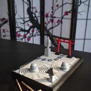 May include: A miniature zen garden set with a red torii gate, a gray pagoda, and a gray stone path. The garden is surrounded by a black frame and includes a rake and a small wooden stick for creating patterns in the sand.