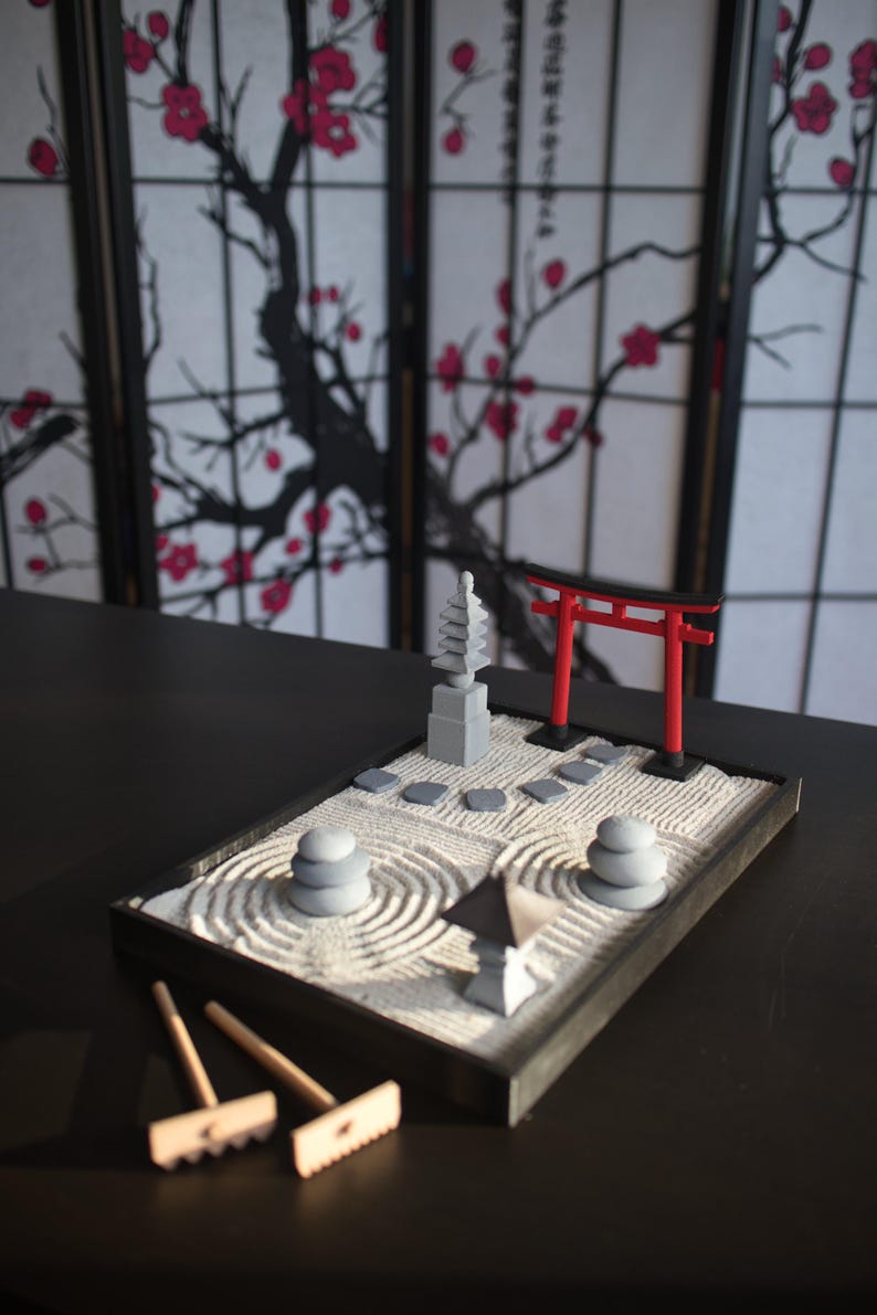 May include: A miniature zen garden set with a red torii gate, a gray pagoda, and a gray stone path. The garden is surrounded by a black frame and includes a rake and a small wooden stick for creating patterns in the sand.