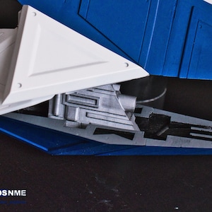 May include: Close-up of a model spaceship with a white and blue paint scheme. The spaceship has a detailed engine and a triangular wing.