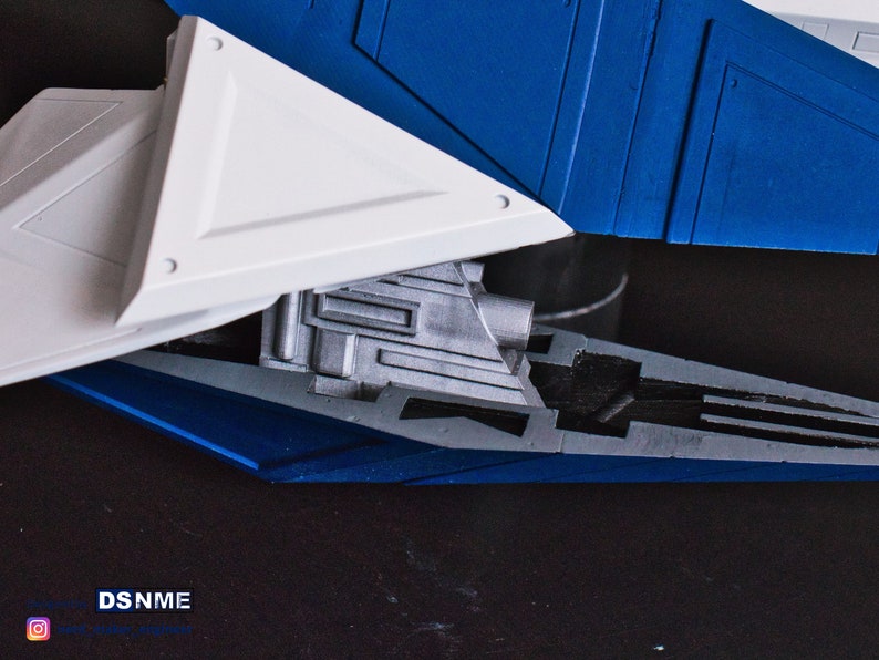 May include: Close-up of a model spaceship with a white and blue paint scheme. The spaceship has a detailed engine and a triangular wing.
