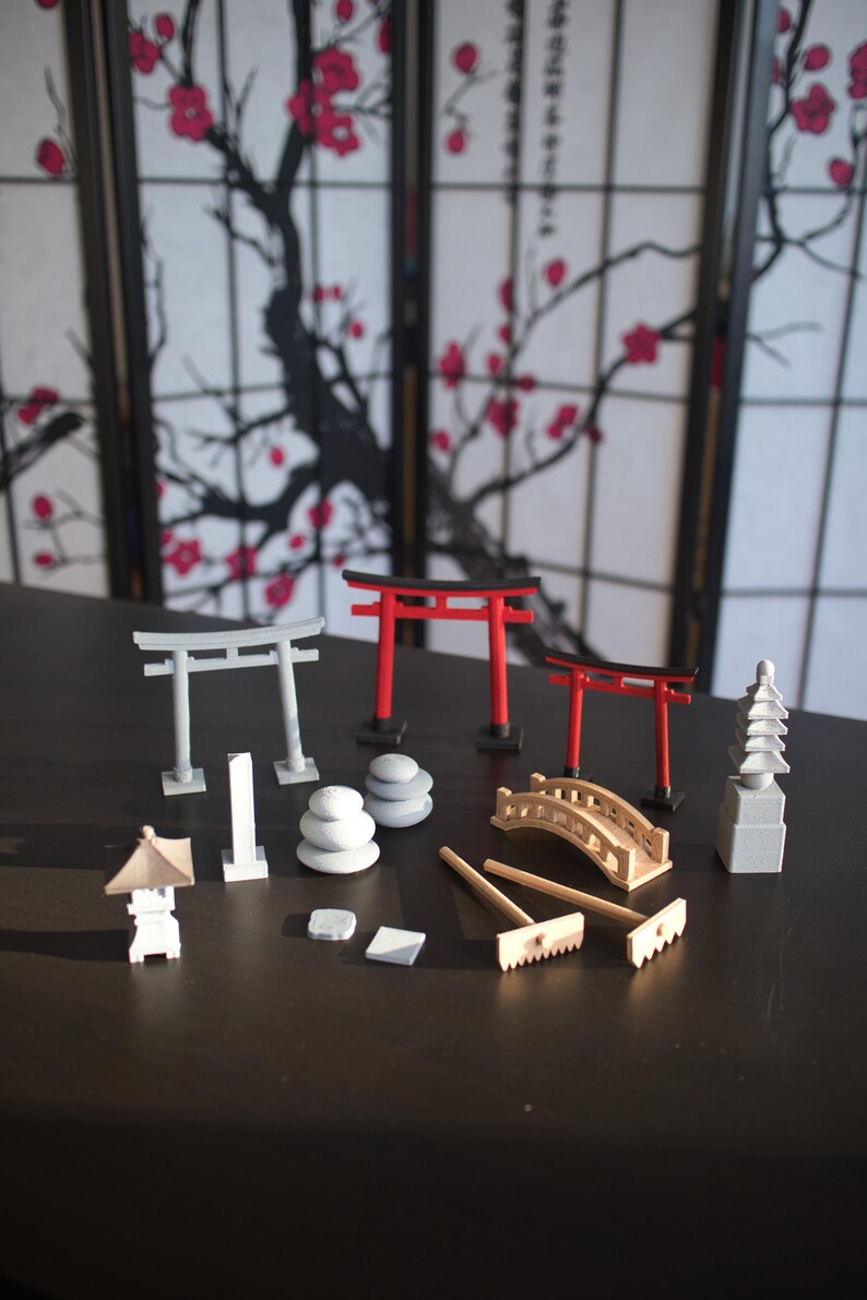 May include: A set of miniature Japanese garden elements, including two red torii gates, two gray torii gates, a gray pagoda, a gray stone lantern, a wooden bridge, a rake, and three stacks of gray stones.