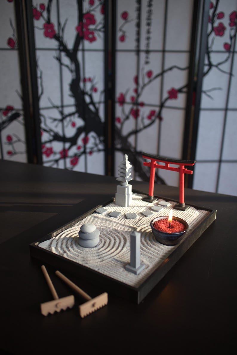 May include: A miniature zen garden with a red torii gate, a small pagoda, a stone path, and a candle in a bowl. The garden is set on a black tray with a wooden rake.