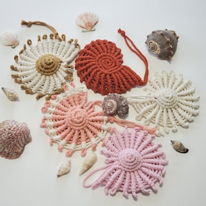May include: Five macrame wall hangings in the shape of a flower with a white background. The wall hangings are in shades of pink, white, and brown.