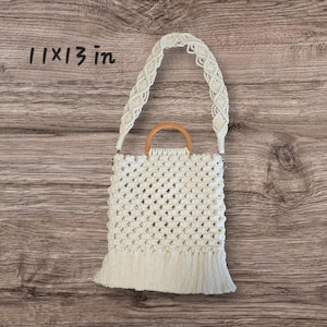 May include: Cream-colored macrame handbag with a wooden handle and a fringed bottom. The bag measures 11 x 13 inches. The woven design creates a diamond pattern. The handle is a light brown color.