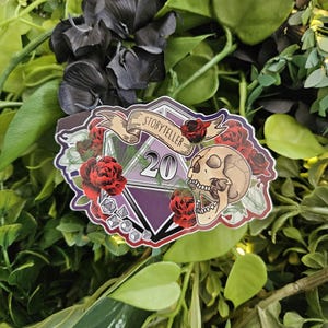 May include: A sticker featuring a D20 die with the number 20, a skull, and red roses. The banner reads "Storyteller". The sticker has a purple and white geometric design.