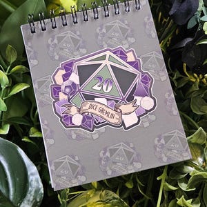 May include: Spiral-bound notebook with a gray cover featuring a repeating pattern of a twenty-sided die. The center design is a large die with the number 20, surrounded by purple gems and a banner that reads "Dice Gremlin."