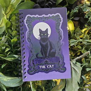 May include: Spiral-bound notebook with a purple cover featuring a black cat illustration. The cat sits in front of a full moon, framed by ornate green vines. The text "THE CAT" is at the bottom. The notebook is surrounded by green foliage.