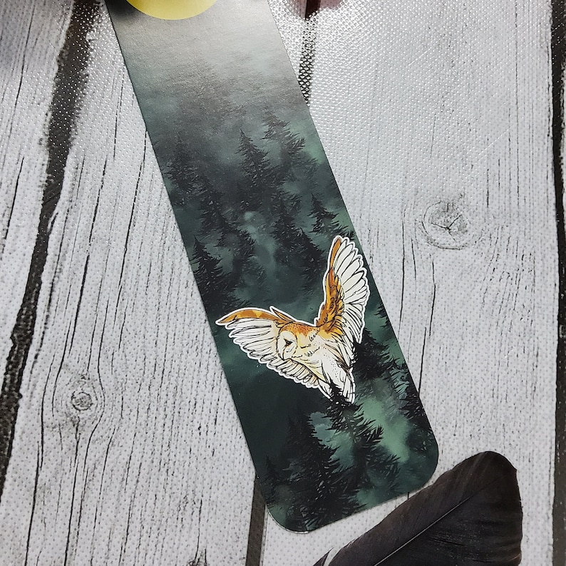 May include: A bookmark featuring a white owl with brown markings flying through a dark green forest with a full moon in the background.