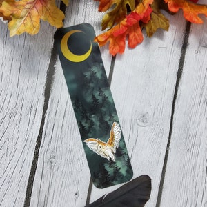May include: A bookmark featuring a dark green forest scene with a white owl flying through the trees. A yellow crescent moon is in the top left corner of the bookmark.