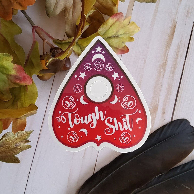 May include: A red and white planchette with the text "Tough Shit" and a black and white skull design. The planchette is shaped like a triangle with a rounded top.