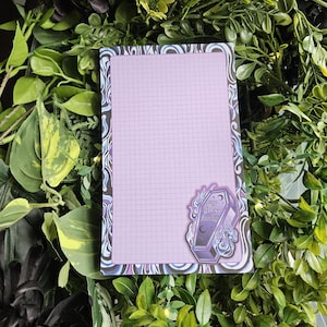 May include: A lavender grid-lined notepad with a decorative border featuring swirling blue and white patterns. A purple coffin-shaped sticker with the text "Witch Dreaming" is affixed to the notepad. The notepad is surrounded by green foliage.