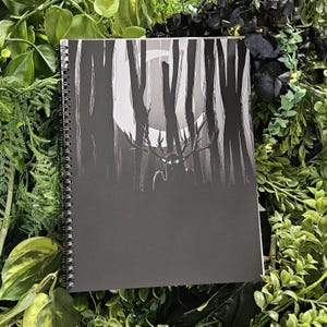 May include: Spiral-bound notebook with a black cover featuring a grayscale illustration of a forest scene. The artwork depicts a figure with antlers in a moonlit forest. The notebook is surrounded by green foliage.