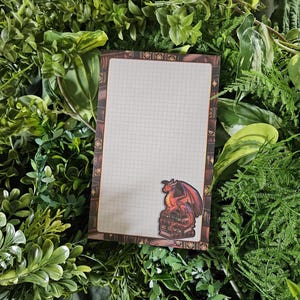 May include: A rectangular notepad featuring a grid pattern on a white background, framed by a border of brown book spines. A small illustration of a red dragon perched atop a stack of books is positioned in the bottom right corner of the page.