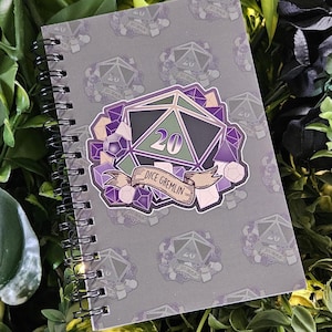 May include: Spiral-bound notebook with a gray cover featuring a repeating pattern of a D20 die. A large D20 die graphic with the text "Dice Gremlin" is centered on the cover. The die is black, green, and purple.