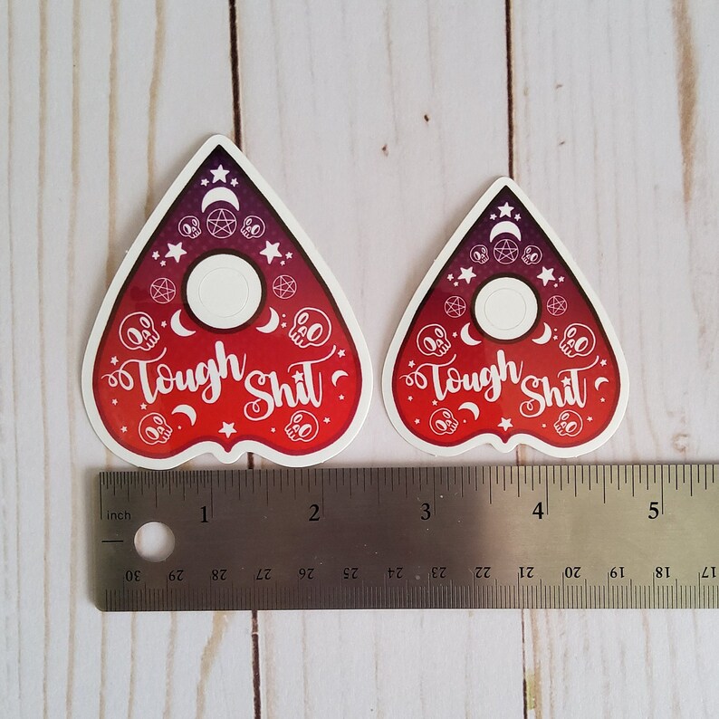 May include: Two red and purple "Tough Shit"  ouija board planchette stickers with white skulls, stars, moons, and pentagrams. The stickers are approximately 2.5 inches tall.