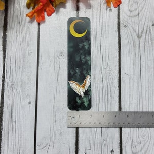May include: A black and green bookmark with a white owl flying in front of a crescent moon and a dark forest. The bookmark is approximately 2 inches tall.