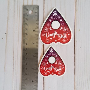 May include: Two red and purple  'Tough Shit'  ouija board-shaped stickers with white text and skull and star designs.