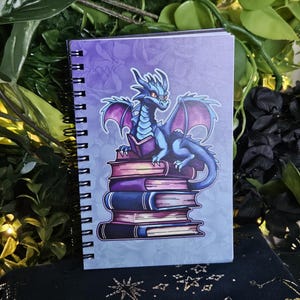 May include: Spiral-bound notebook featuring a blue dragon perched atop a stack of books. The dragon has purple wings and red eyes, set against a purple background. The notebook's cover has a fantasy theme.