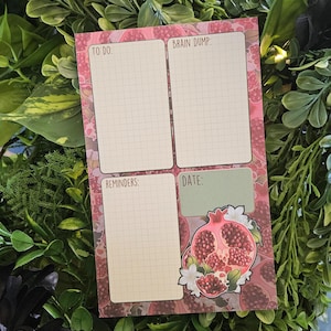 May include: A pink and white notepad with four sections labeled "TO DO:", "BRAIN DUMP:", "REMINDERS:", and "DATE:". The notepad features a pomegranate illustration with white flowers and a grid pattern.