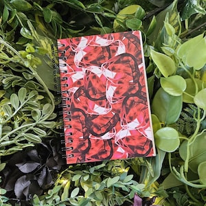 May include: A spiral-bound notebook with a vibrant cover design featuring a repeating pattern of red and white geometric shapes. The notebook is surrounded by lush green foliage, creating a visually appealing contrast.