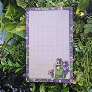 May include: A rectangular notepad with a grid pattern and a purple border adorned with floral designs. A decorative illustration of a potion bottle with a skull and crossbones is in the lower right corner. The notepad is surrounded by green foliage.